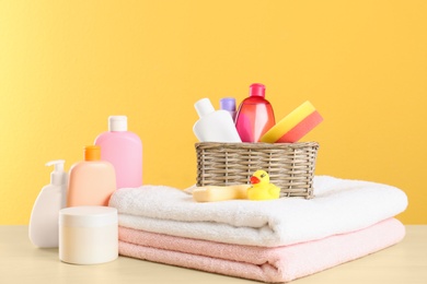 Photo of Wicker basket with baby cosmetic products, toy and towels on table against color background