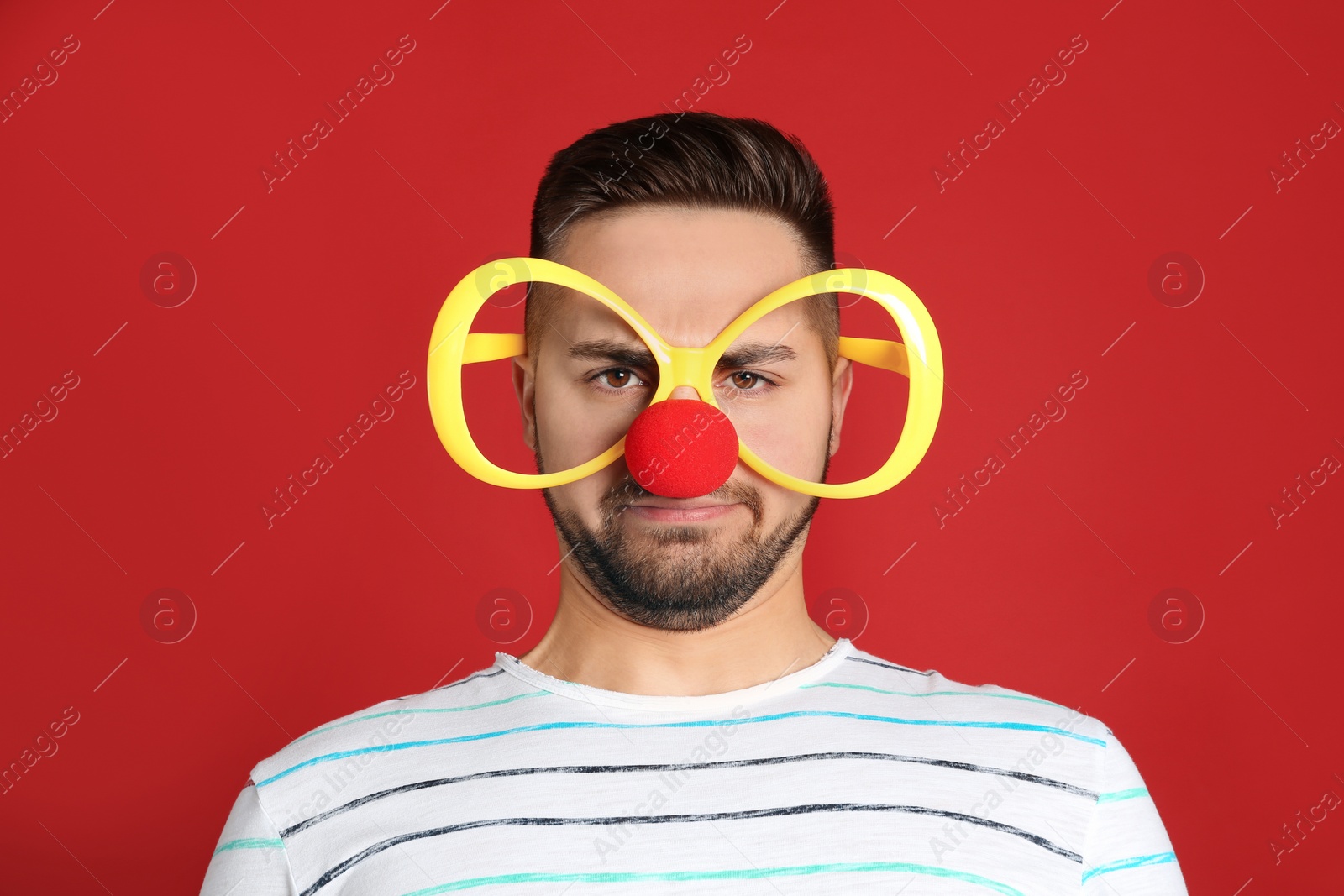 Photo of Emotional young man with party glasses and clown nose on red background. April fool's day