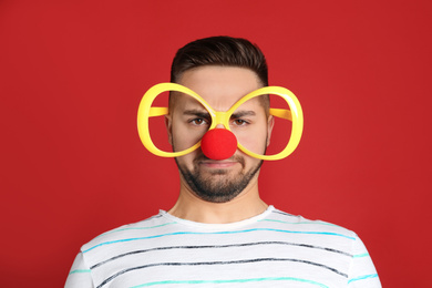 Photo of Emotional young man with party glasses and clown nose on red background. April fool's day
