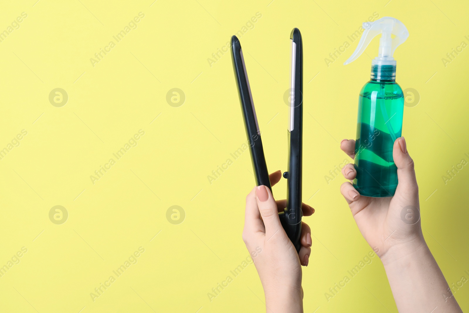 Photo of Woman holding spray bottle with thermal protection and stylish straightener on pale yellow background, closeup. Space for text