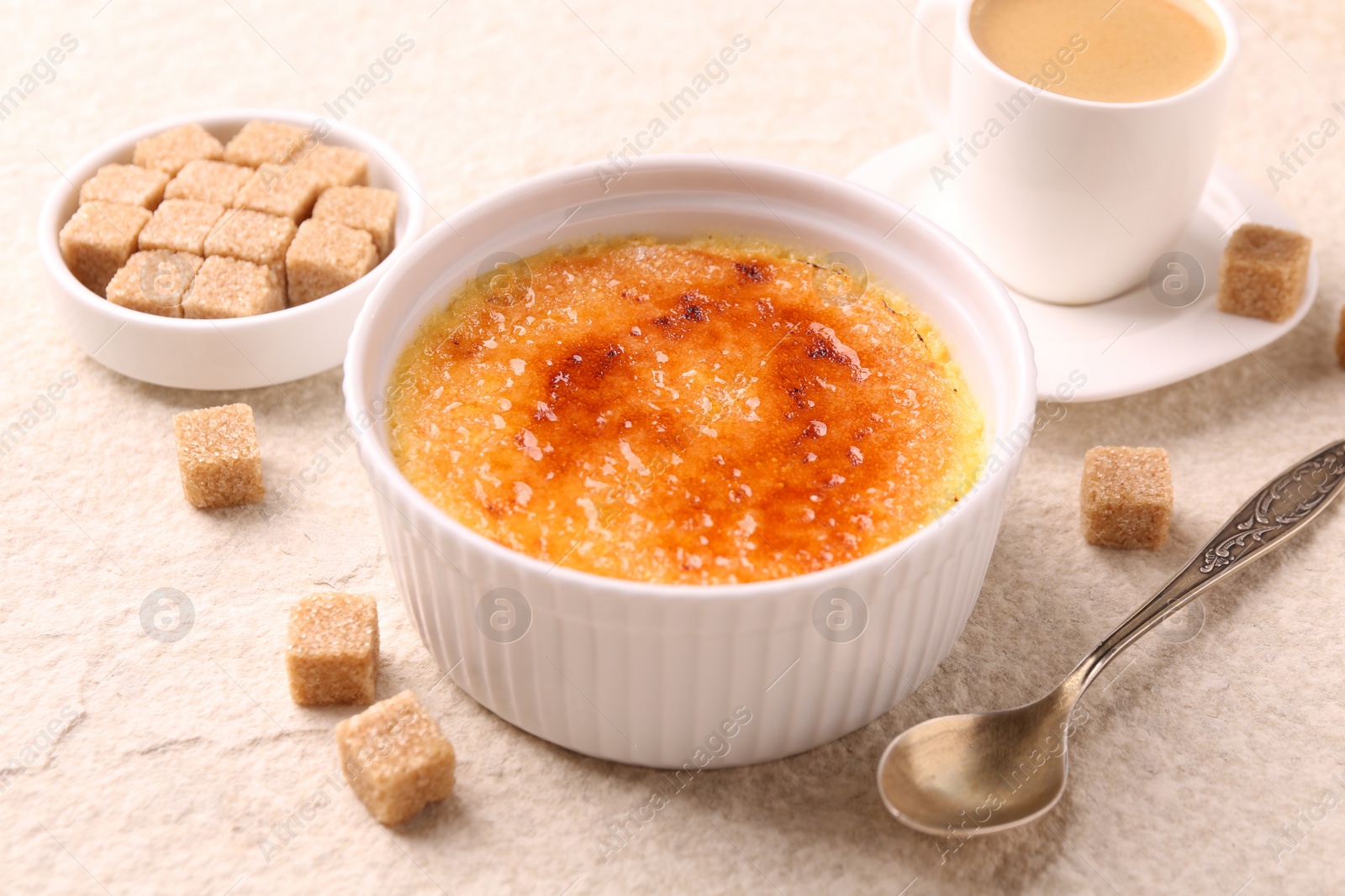 Photo of Delicious creme brulee in bowl, sugar cubes, coffee and spoon on light textured table, closeup