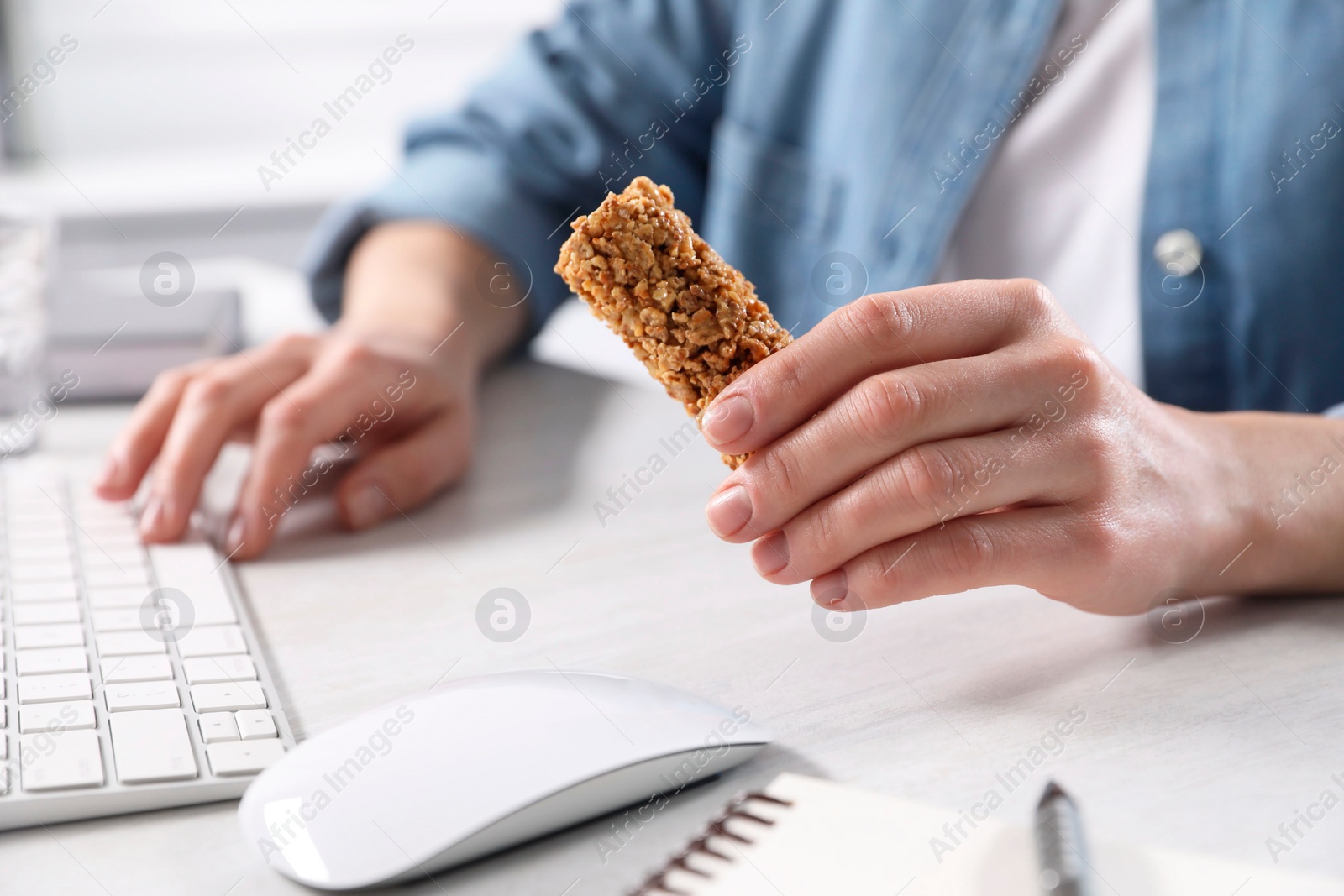 Photo of Woman holding tasty granola bar working with computer at light table in office, closeup