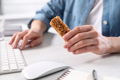 Woman holding tasty granola bar working with computer at light table in office, closeup