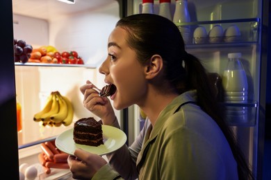 Young woman eating cake near refrigerator in kitchen at night. Bad habit