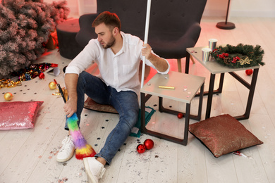 Man with dusting brush and mop suffering from hangover in messy room after New Year party