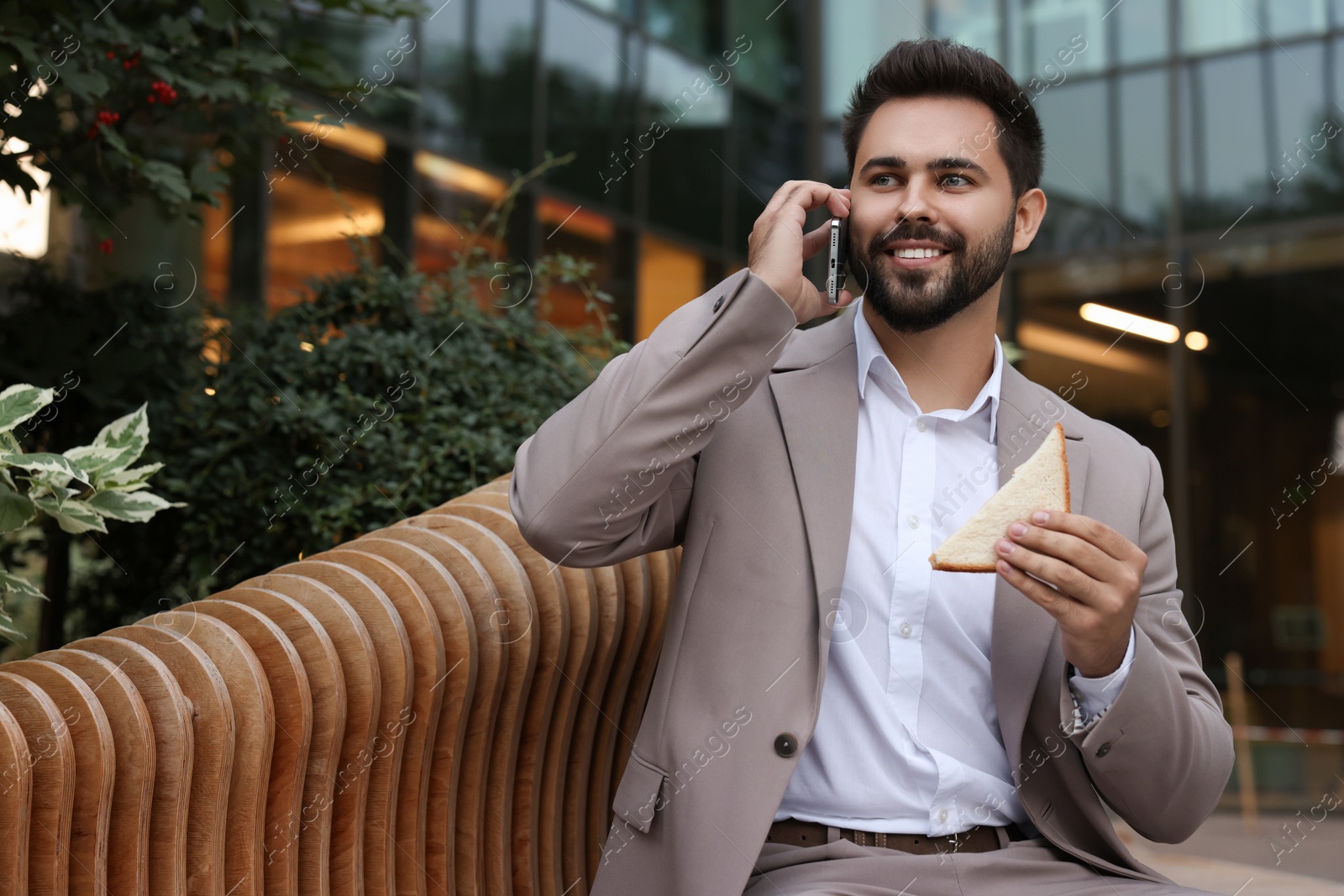 Photo of Lunch time. Young businessman with sandwich talking on smartphone on bench outdoors