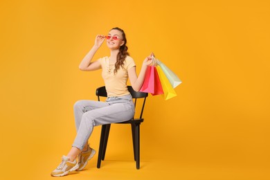 Happy woman in stylish sunglasses holding colorful shopping bags on chair against orange background