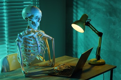 Photo of Waiting concept. Human skeleton sitting at wooden table with laptop indoors