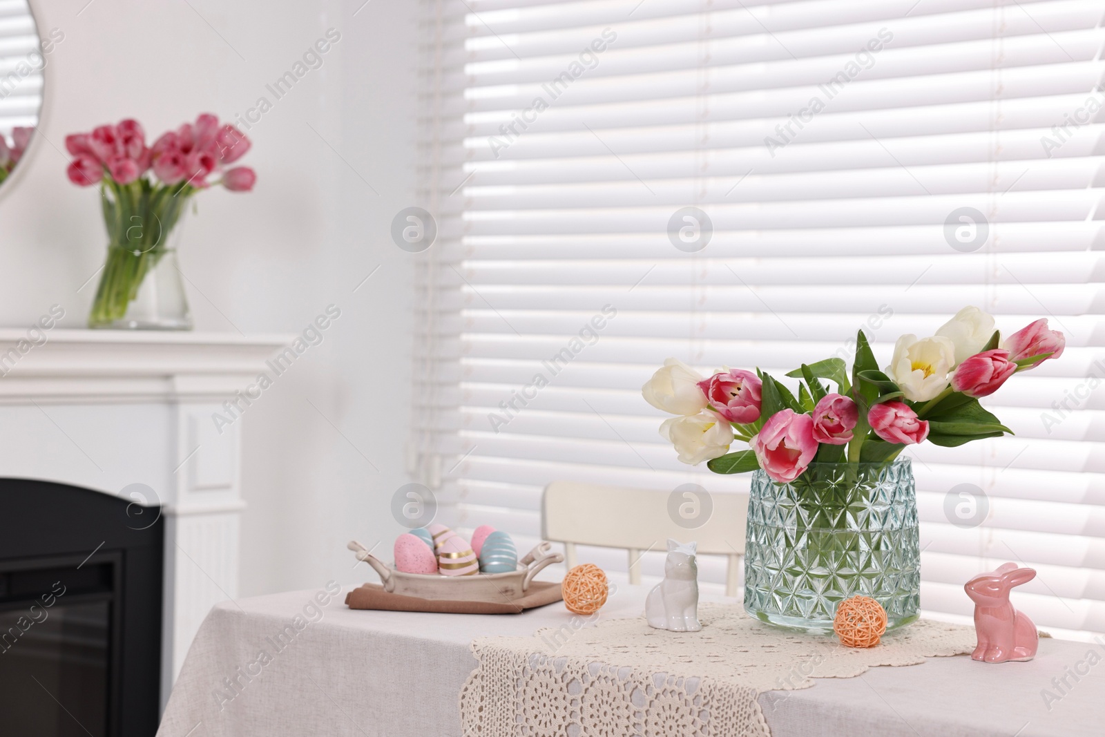 Photo of Easter decorations. Bouquet of tulips in vase, painted eggs and bunny figures on white table at home