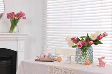 Photo of Easter decorations. Bouquet of tulips in vase, painted eggs and bunny figures on white table at home