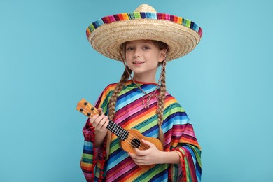 Cute girl in Mexican sombrero hat and poncho playing ukulele on light blue background