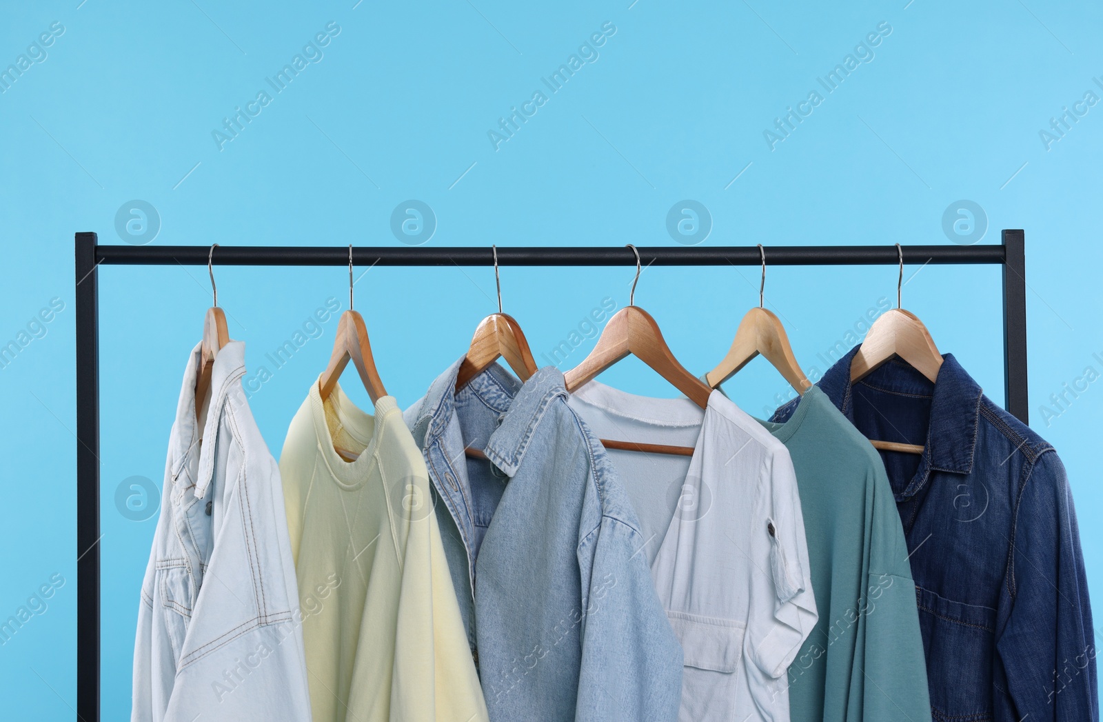 Photo of Rack with stylish clothes on wooden hangers against light blue background