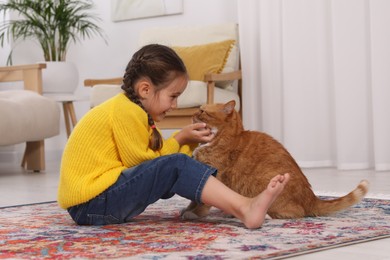 Photo of Happy little girl playing with cute ginger cat on carpet at home