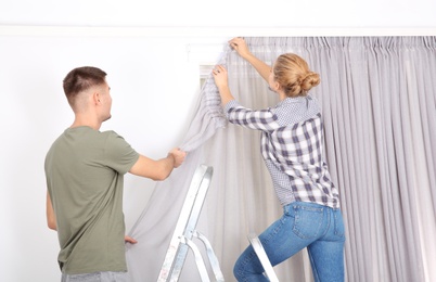 Young couple hanging window curtain indoors. Interior decor element