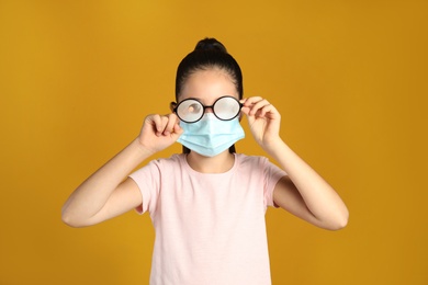 Photo of Little girl wiping foggy glasses caused by wearing medical face mask on yellow background. Protective measure during coronavirus pandemic