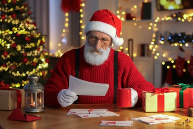 Photo of Santa Claus reading letter and drinking hot beverage at his workplace in room decorated for Christmas