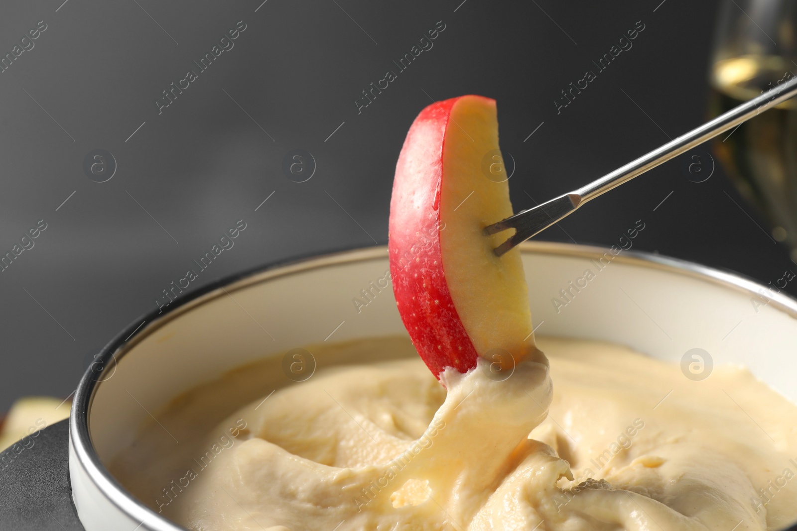 Photo of Dipping piece of apple into fondue pot with melted cheese on grey background, closeup