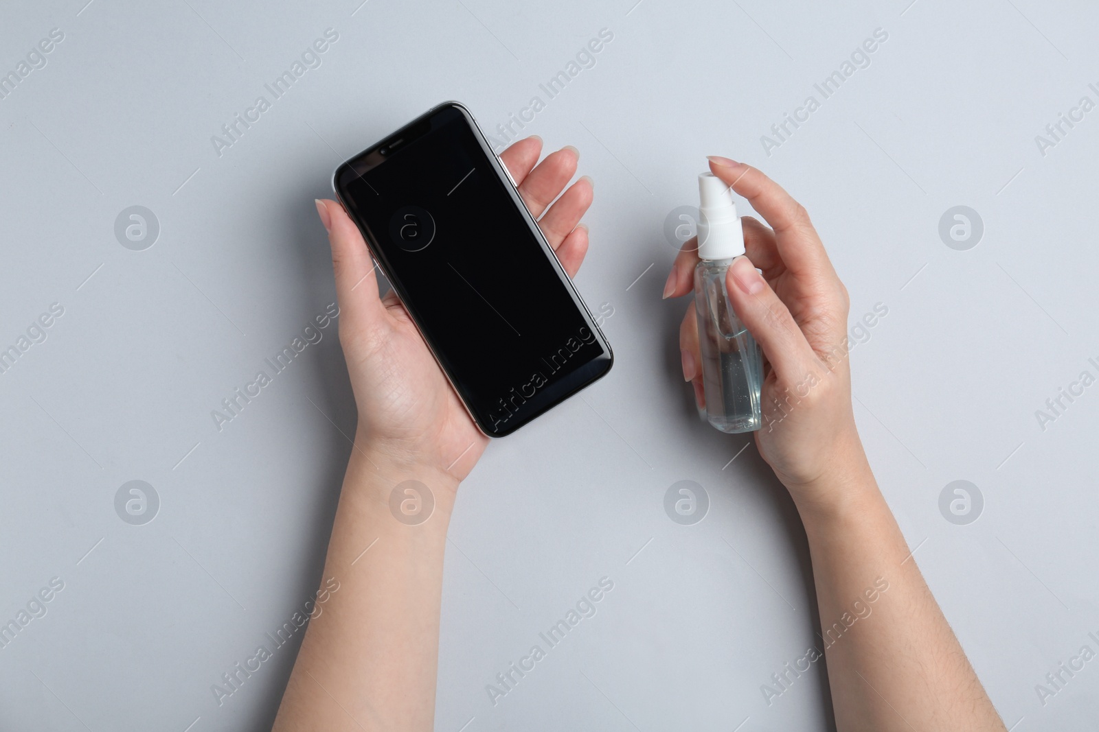 Photo of Woman spraying antiseptic onto smartphone on light grey background, top view