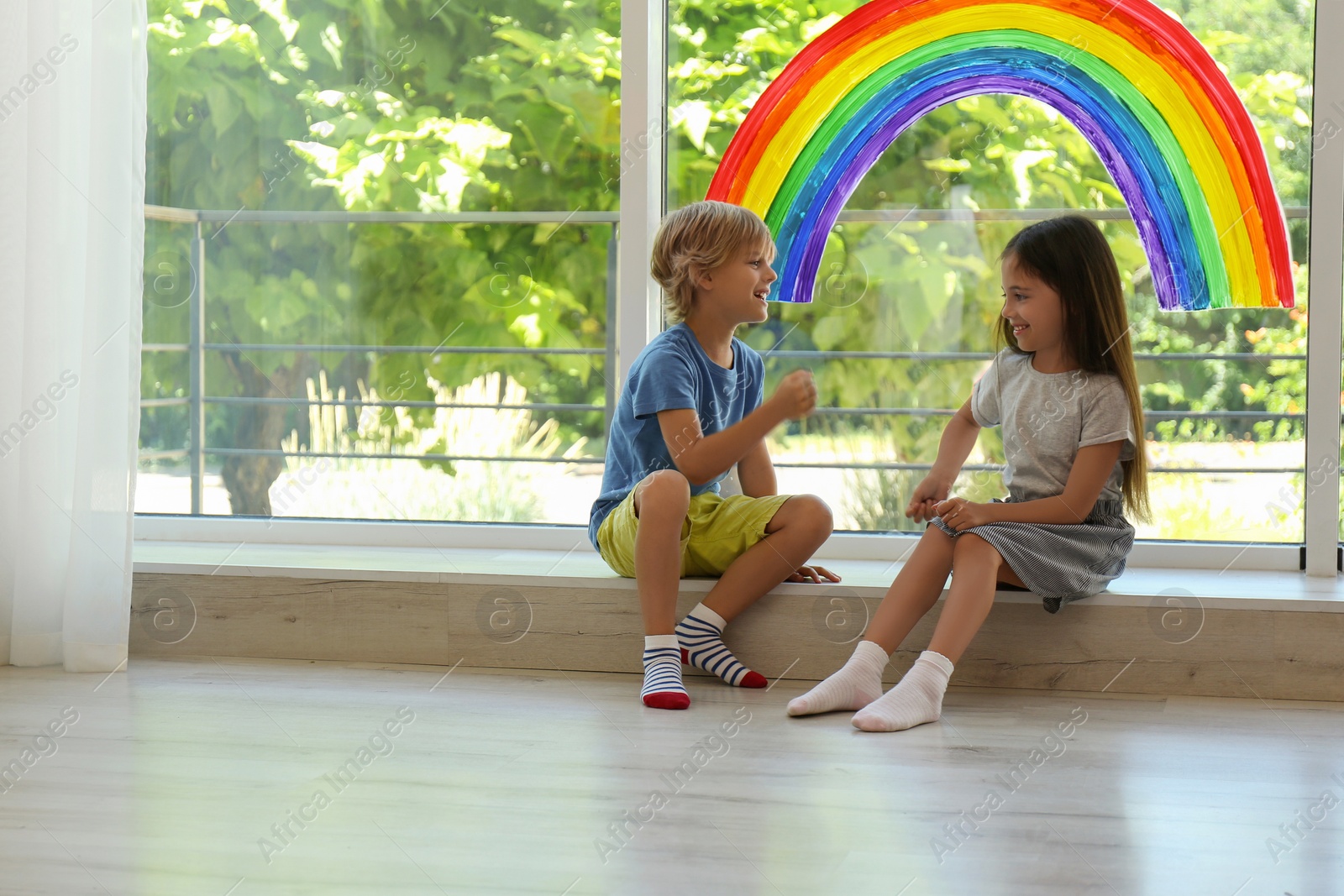 Photo of Little children playing near rainbow painting on window indoors. Stay at home concept