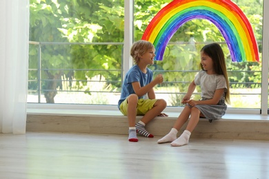 Photo of Little children playing near rainbow painting on window indoors. Stay at home concept