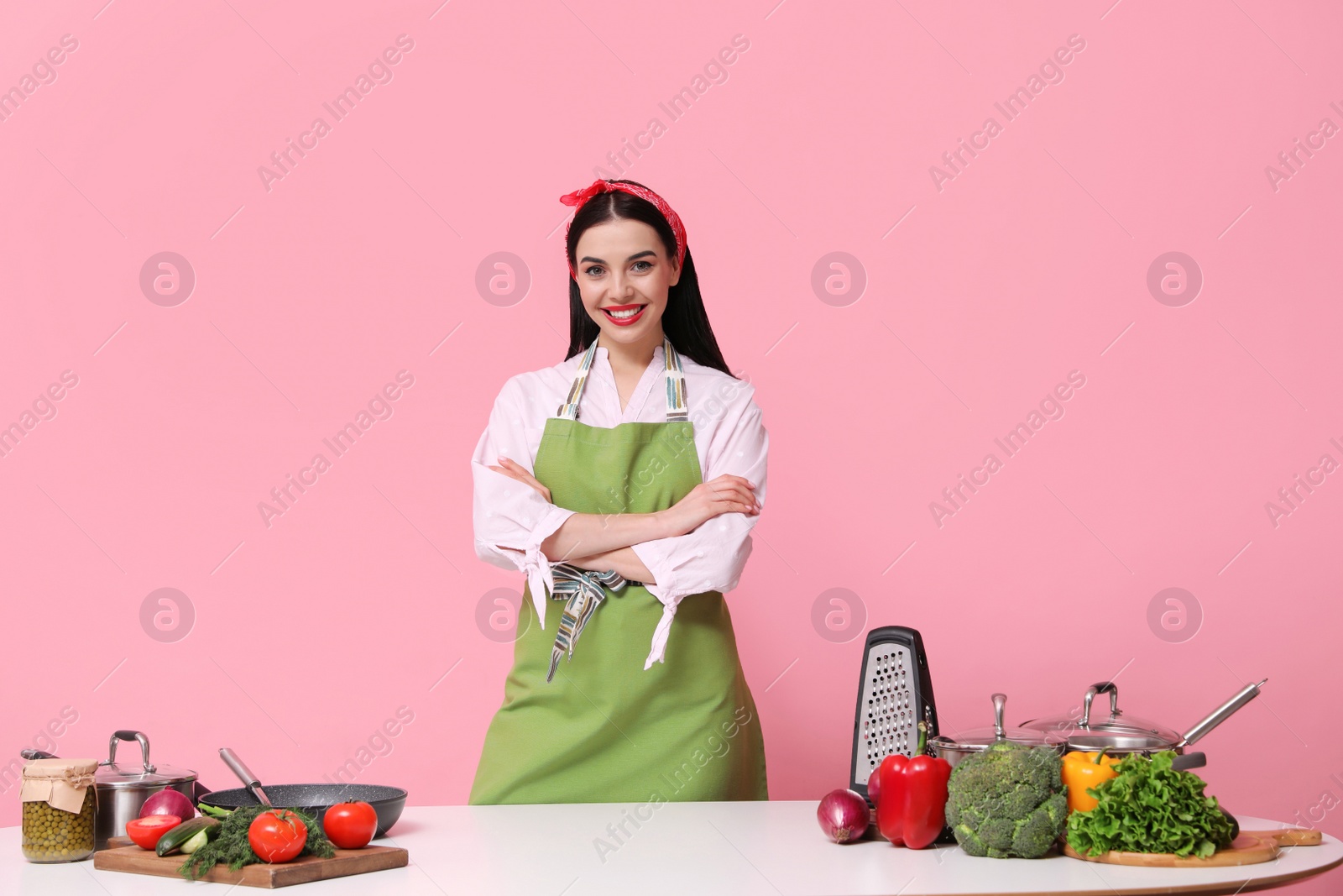 Photo of Young housewife at white table with utensils and products on pink background