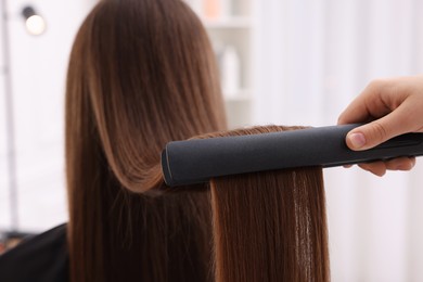 Photo of Hairdresser straightening woman's hair with flat iron indoors, closeup