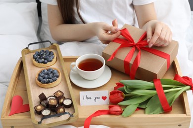 Photo of Tasty breakfast served in bed. Woman with gift box, desserts, tea, flowers and I Love You card at home, closeup