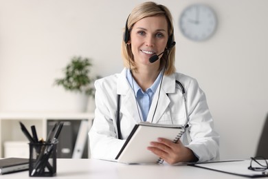 Photo of Smiling doctor in headphones with notebook having online consultation at table indoors