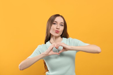 Beautiful young woman blowing kiss and making heart with hands on yellow background