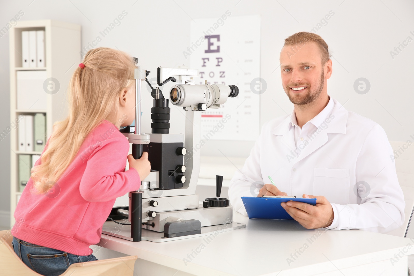 Photo of Children's doctor examining little girl with ophthalmic equipment in clinic