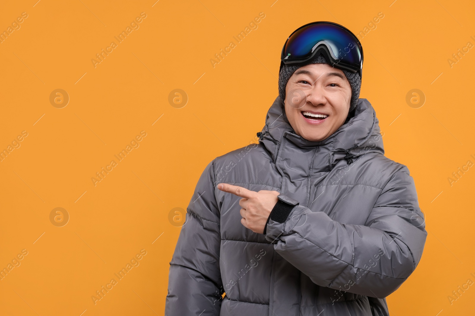 Photo of Winter sports. Happy man with ski goggles pointing at something on orange background. Space for text