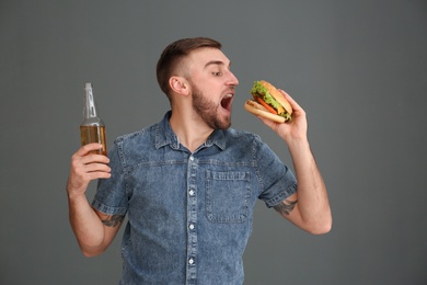 Young man with beer eating tasty burger on grey background