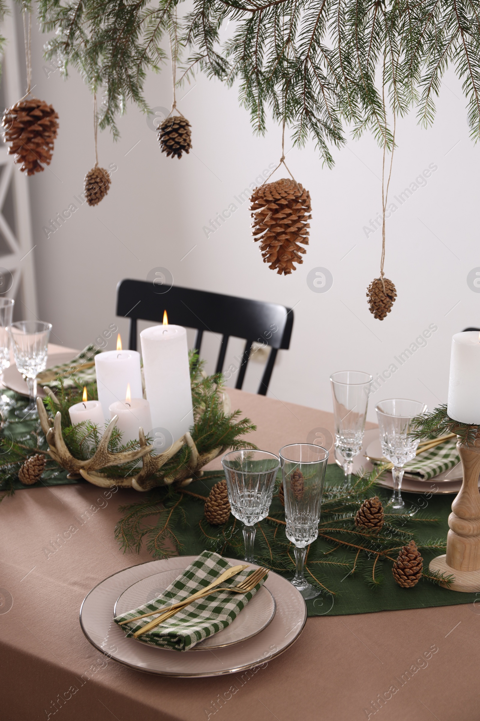 Photo of Christmas celebration. Cones hanging from fir tree branch over table with burning candles and tableware