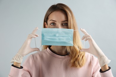 Young woman in medical gloves putting on protective mask against grey background