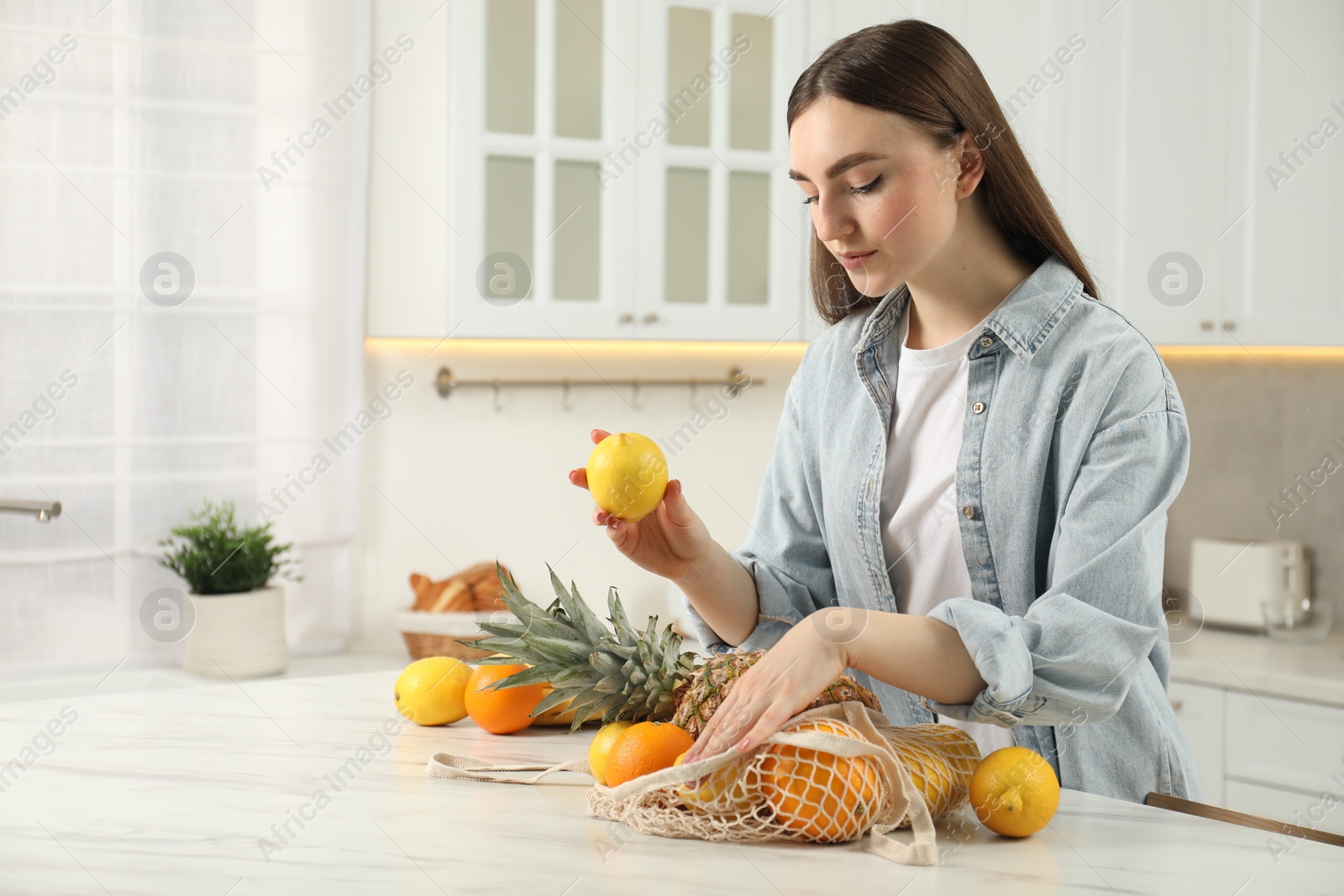 Photo of Woman with lemon and string bag of fresh fruits at light marble table in kitchen