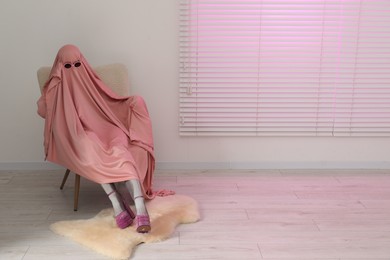 Glamorous ghost. Woman in pink sheet and high heel shoes on armchair indoors, space for text