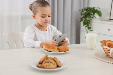 Photo of Little girl using smartphone while having breakfast at table indoors. Internet addiction