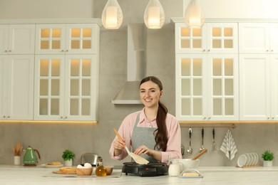 Photo of Happy woman cooking delicious crepe on electric maker at white marble table in kitchen