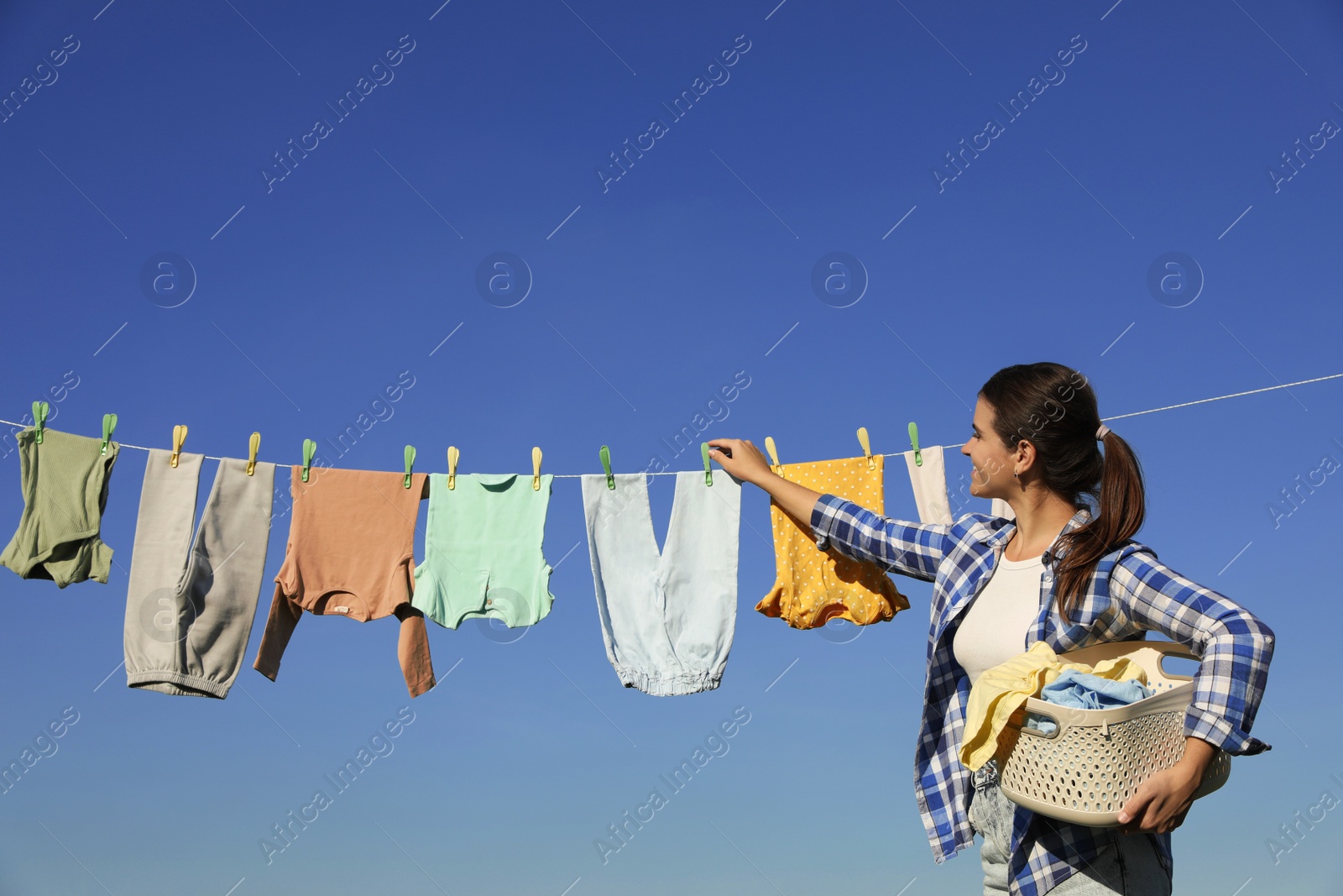 Photo of Smiling woman hanging baby clothes with clothespins on washing line for drying against blue sky outdoors