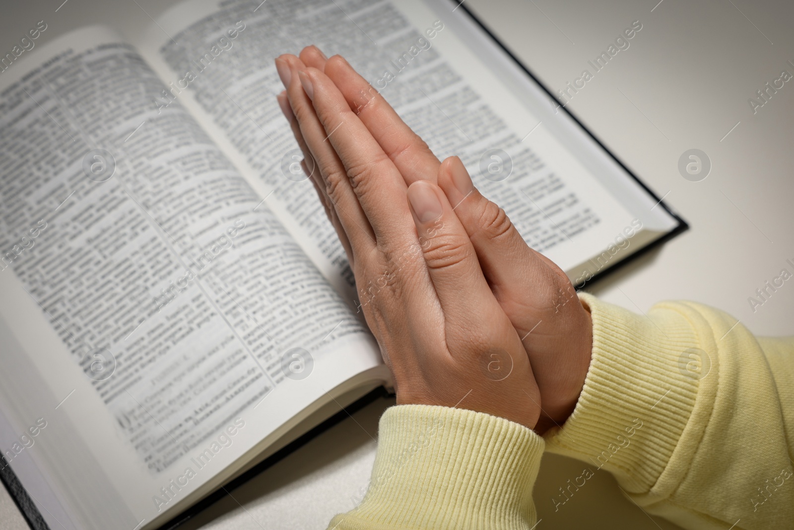 Photo of Woman holding hands clasped while praying over Bible at white table, closeup