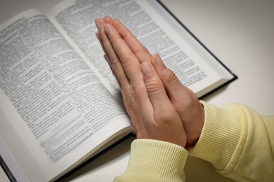 Photo of Woman holding hands clasped while praying over Bible at white table, closeup