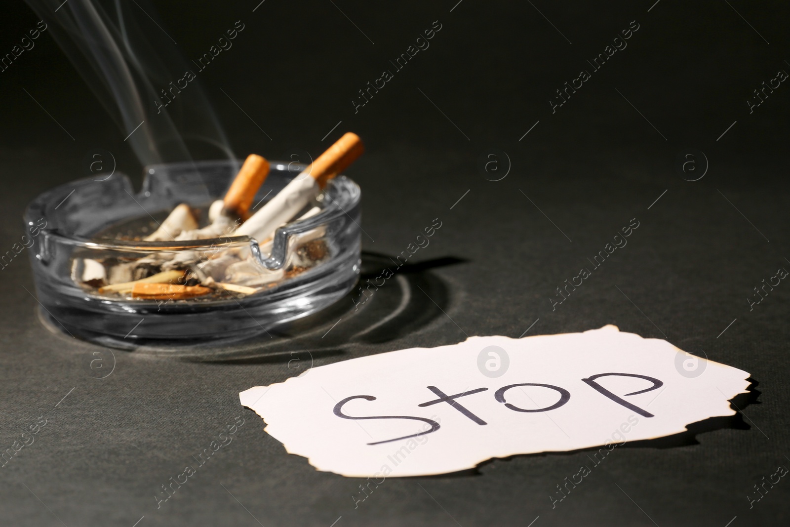 Photo of Ashtray with burnt cigarettes and word Stop written on paper on black table, closeup. No smoking concept