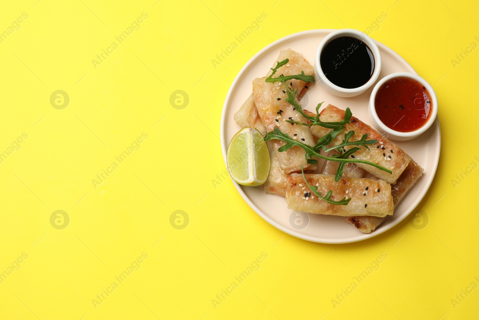 Photo of Tasty fried spring rolls with arugula, lime and sauces on yellow table, top view. Space for text