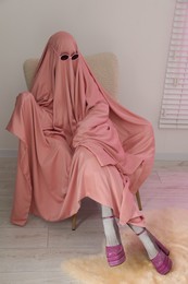 Glamorous ghost. Woman in pink sheet and high heel shoes on armchair indoors