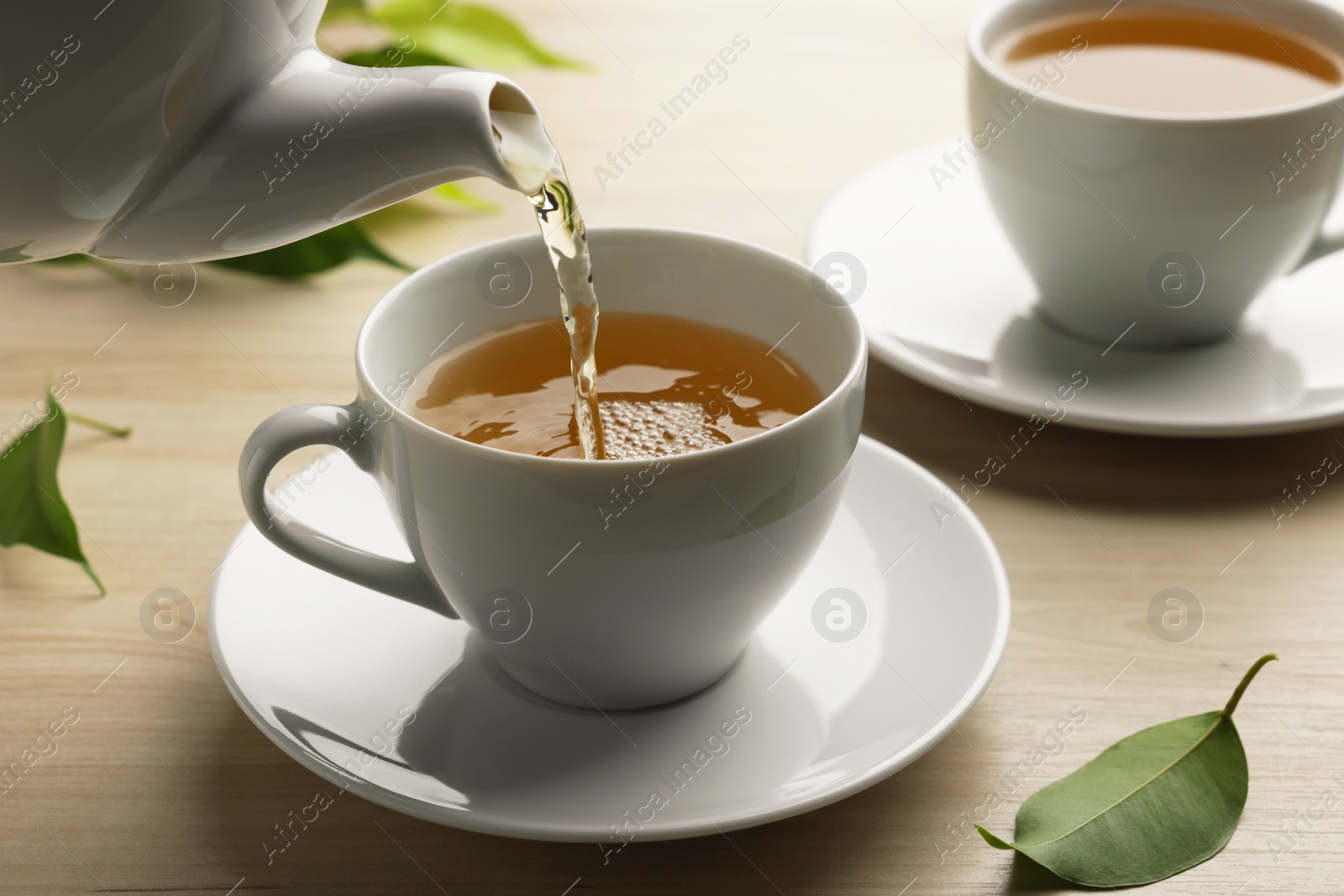 Photo of Pouring green tea into white cup with saucer and leaves on wooden table, closeup