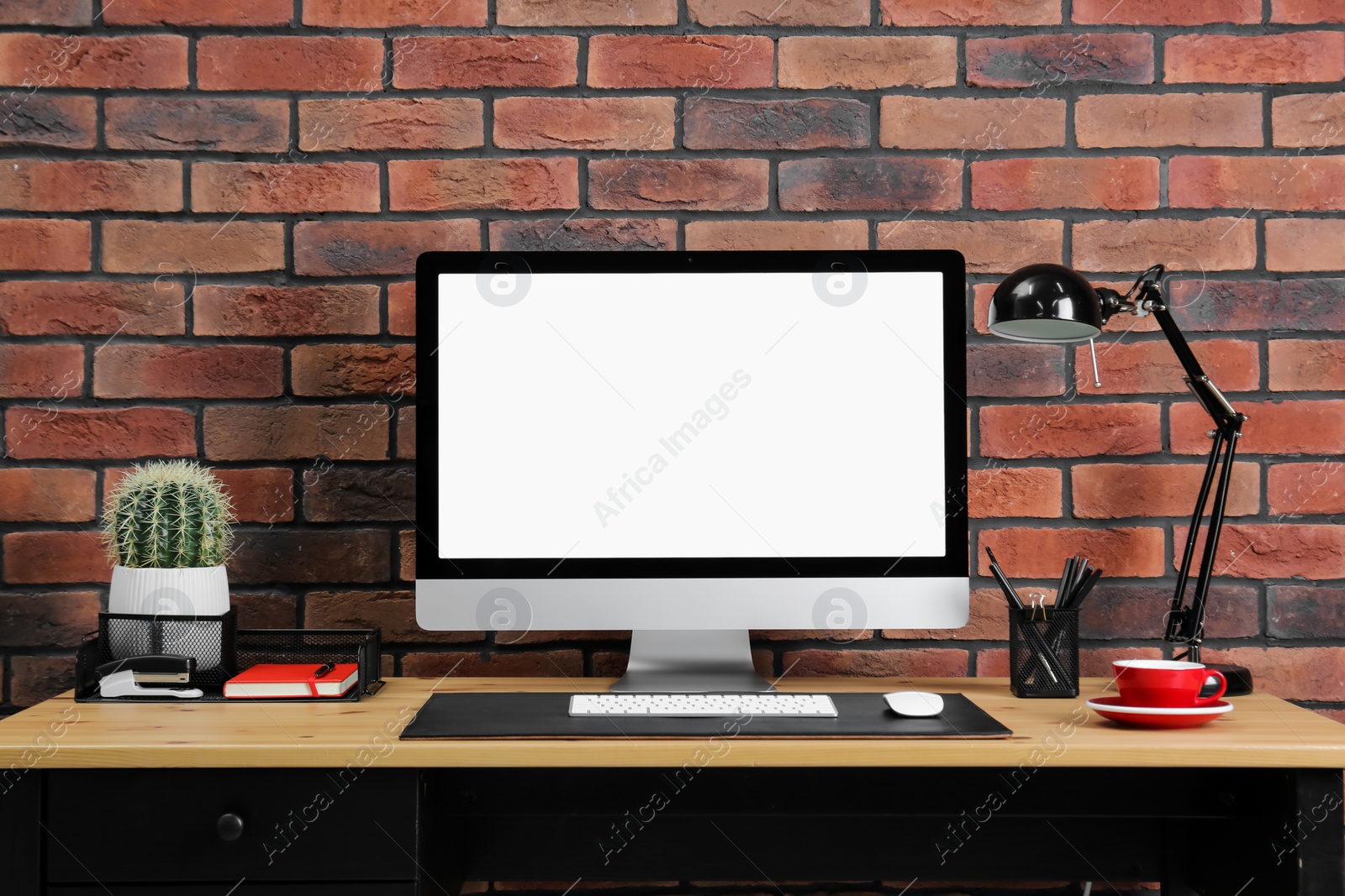 Photo of Stylish workplace with computer, lamp and stationery on wooden table near brick wall