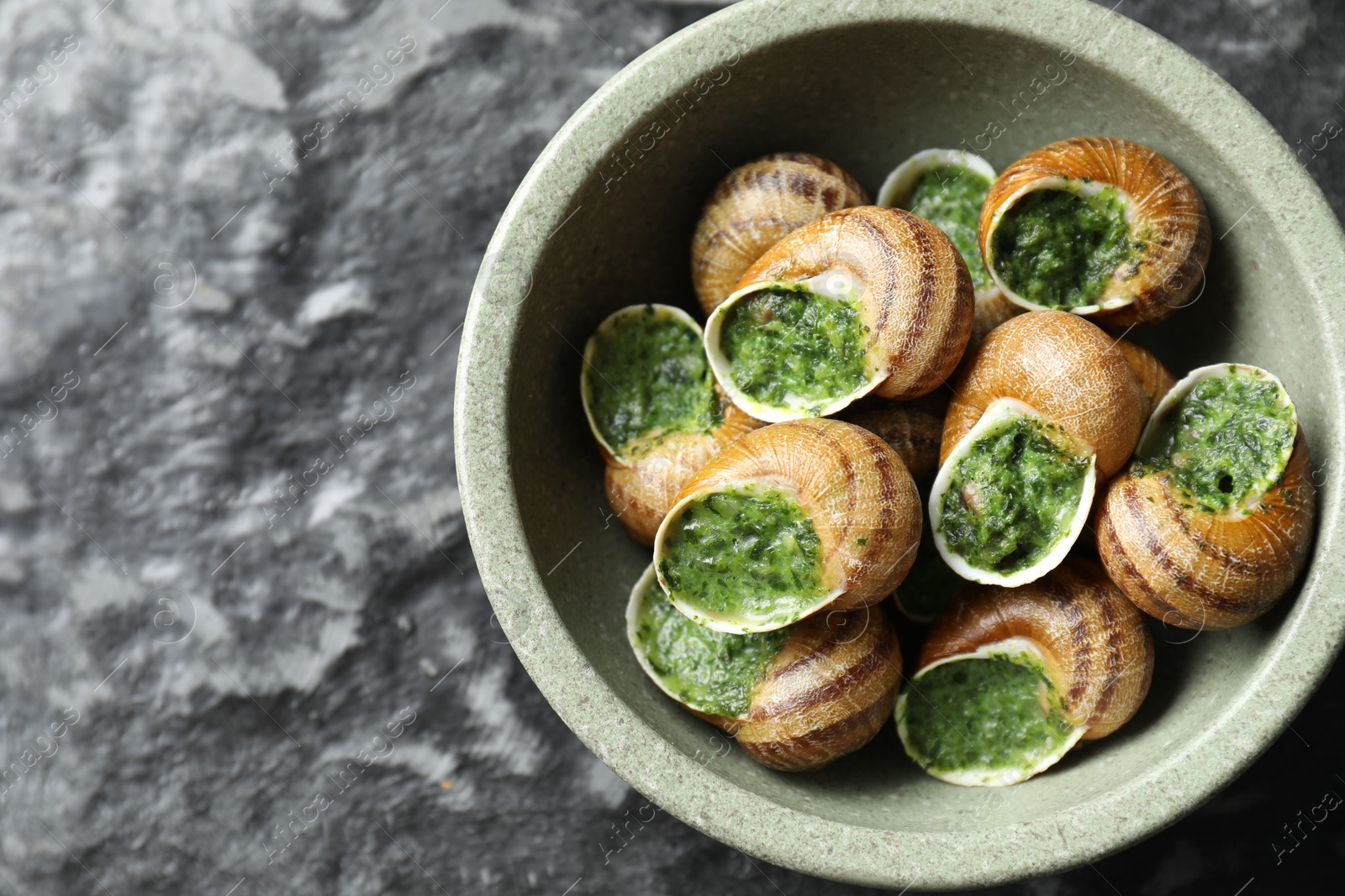 Photo of Delicious cooked snails in bowl on grey textured table, top view. Space for text