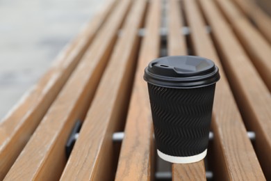 Black disposable paper cup with plastic lid on wooden bench outdoors. Space for text