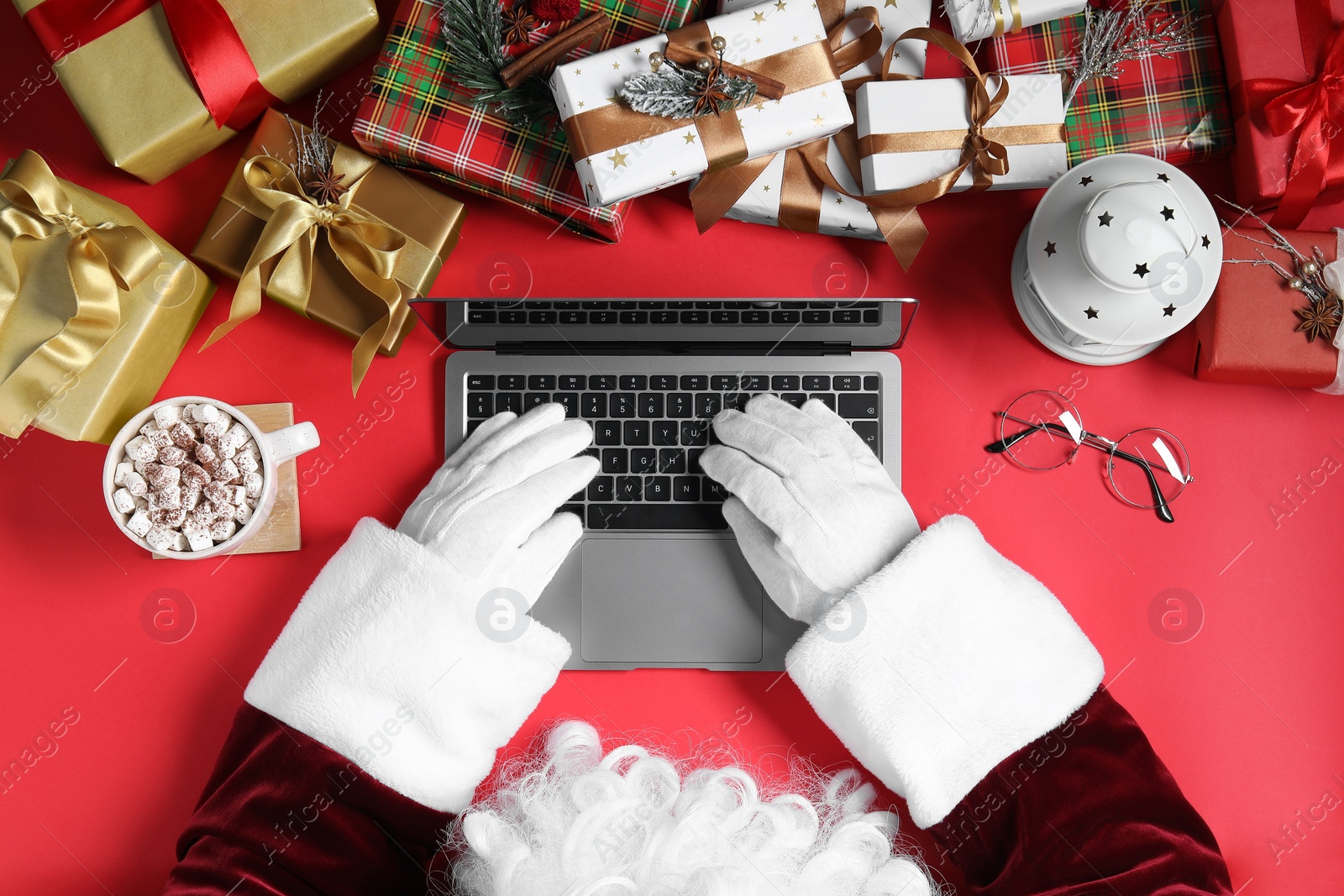 Photo of Santa Claus using laptop, closeup. Gift boxes, cup of drink and Christmas decor on red background, top view