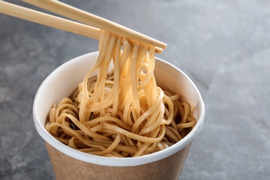 Photo of Paper cup of instant noodles with sauce and chopsticks on grey background, closeup. Space for text